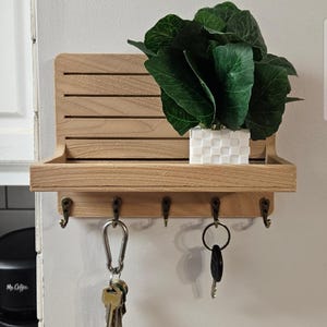 May include: A wooden key holder with a shelf and five hooks. A small white square patterned pot with a green plant sits on the shelf. Keys hang from two of the hooks. The key holder is mounted on a white wall.