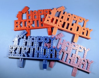 May include: Three cake toppers in different colors, orange, blue, and pink, spelling out "Happy Birthday" in a Lego-style font. Each topper has a small Lego figure on top.