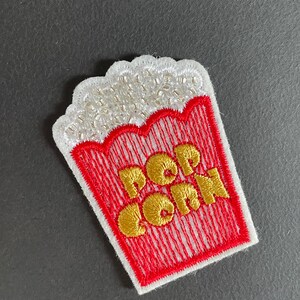 Może przedstawiać: Haftowana naszywka w kształcie pojemnika na popcorn. Naszywka ma czerwono-biały wzór ze złotymi literami "POP CORN". Górna część naszywki jest biała z małymi, srebrnymi koralikami.