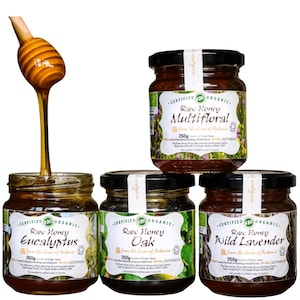 May include: Four jars of raw organic honey, including eucalyptus, oak, wild lavender, and multifloral varieties. Each jar is 250g and has a black lid with a white label. Honey drips from a wooden honey dipper into one jar.