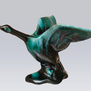 May include: A ceramic sculpture of a goose in flight, with outstretched wings. The goose is primarily teal and black, with a bronze-colored beak. The sculpture has a glossy finish and is mounted on a small base.