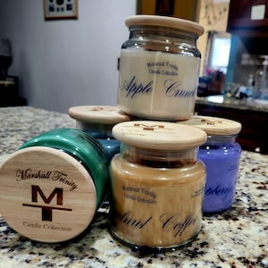 May include: Four glass candle jars with wooden lids. The candles are labeled with the text "Marshall Trinity Candle Collection". The candle scents are "Apple Crunch", "Walnut Coffee", "Raspberry", and an unreadable scent.