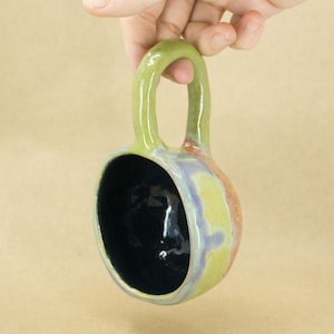 May include: A small, green, ceramic mug with a handle. The mug has a dark blue interior and a colorful glaze with shades of purple, yellow, and orange.