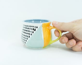 Può includere: Una tazza in ceramica con un design geometrico in nero, bianco, azzurro e arancione. La tazza ha una forma arrotondata e un manico.