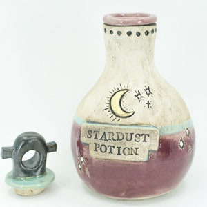 Może przedstawiać: Ceramiczną butelkę z korkowym korkiem, ozdobioną księżycem i gwiazdami. Butelka jest oznaczona etykietą "Stardust Potion" i ma białą, niebieską i fioletową glazurę.