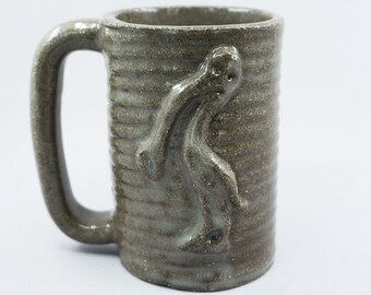 May include: A brown ceramic mug with a textured, striped design. A three-dimensional figure is molded into the side of the mug.