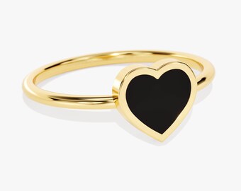 May include: A gold ring featuring a smooth, polished band and a central heart-shaped setting. The heart is inlaid with a solid, flat black material, creating a distinct contrast against the bright, reflective yellow gold metal of the ring.