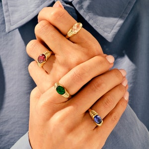 May include: Four gold rings with different colored gemstones. The rings have a twisted band design. The gemstones are a light pink, a dark red, a dark green, and a dark blue.