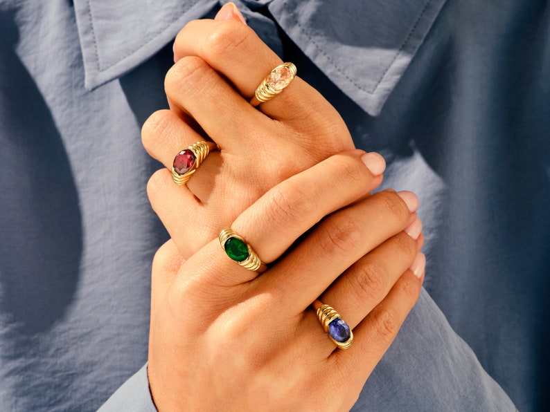 May include: Four gold rings with different colored gemstones. The rings have a twisted band design. The gemstones are a light pink, a dark red, a dark green, and a dark blue.