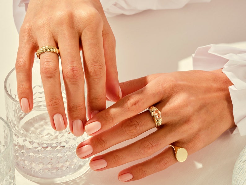 May include: Close-up of two hands wearing gold rings. The rings are a thick, twisted band and a ring with a clear stone set in a gold band. The hands are resting on a glass of water.