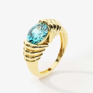 May include: A gold ring with a large, oval-shaped, light blue gemstone set in the center. The band of the ring is intricately designed with multiple layers of gold.