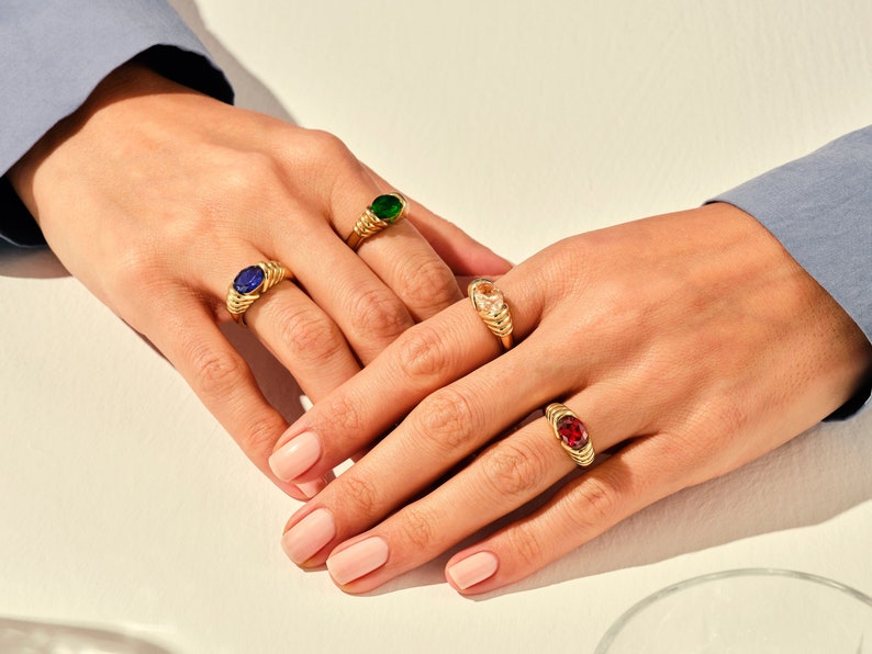 May include: Four gold rings with different colored gemstones. The rings feature a twisted band design. The gemstones are blue, green, red, and light orange.