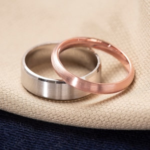 May include: Two wedding bands, one silver and one rose gold, are stacked on top of each other on a beige fabric background.