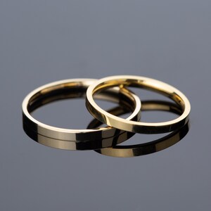 May include: Two gold wedding bands, one slightly larger than the other, are stacked on top of each other on a reflective surface.
