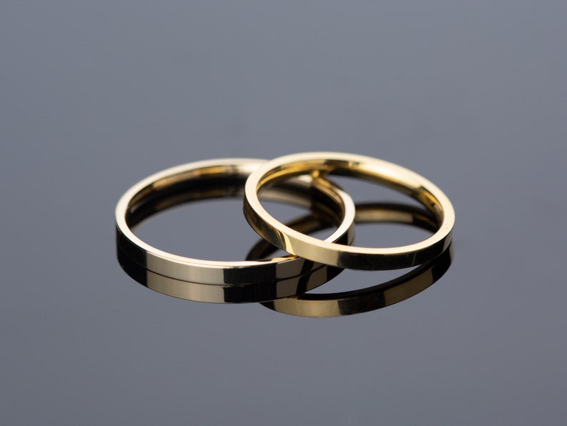 May include: Two gold wedding bands, one slightly larger than the other, are stacked on top of each other on a reflective surface.