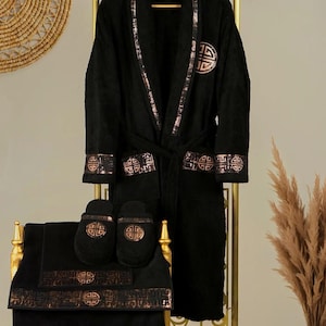 Op de afbeelding: Zwarte badjas met gouden bies en bijpassende slippers en handdoeken. De badjas heeft een traditioneel Chinees symbool op de voorkant en de handdoeken en slippers hebben hetzelfde symbool.