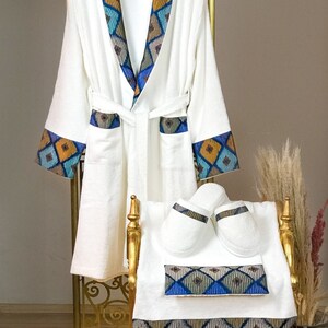 Op de afbeelding: Witte badjas met een blauwe, gele en bruine geometrische rand. De badjas heeft een bijpassende badhanddoek en slippers.