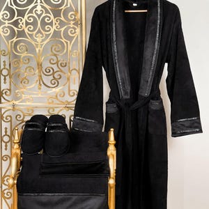 Op de afbeelding: Zwarte badjas met een satijnen rand, bijpassende slippers en een handdoek. De badjas heeft een shawlkraag en een strikceintuur.
