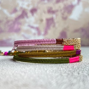 以下が含まれることがあります： 4つのブレスレットが積み重ねられています。一番上のブレスレットは金色のチェーン、2番目は薄いピンク色の編み紐、3番目は薄いベージュ色の編み紐、一番下のブレスレットは濃い緑色の編み紐です。すべてのブレスレットには明るいピンク色のストライプがあります。