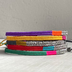 May include: A stack of colourful bracelets with different patterns and textures. The bracelets are made of woven string and metal chain. The colours include purple, yellow, grey, red, turquoise, and pink.