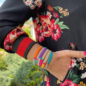 May include: A close-up of a person's wrist wearing a stack of colorful beaded bracelets. The bracelets are made of different colors, including pink, blue, green, and gold. The person is wearing a black floral jacket with red trim.
