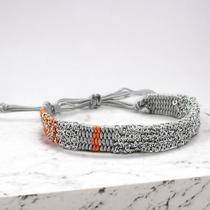 Pode incluir: Um bracelete trançado prateado e laranja com fecho de nó. O bracelete é feito de um cordão tecido com uma corrente prateada entrelaçada.