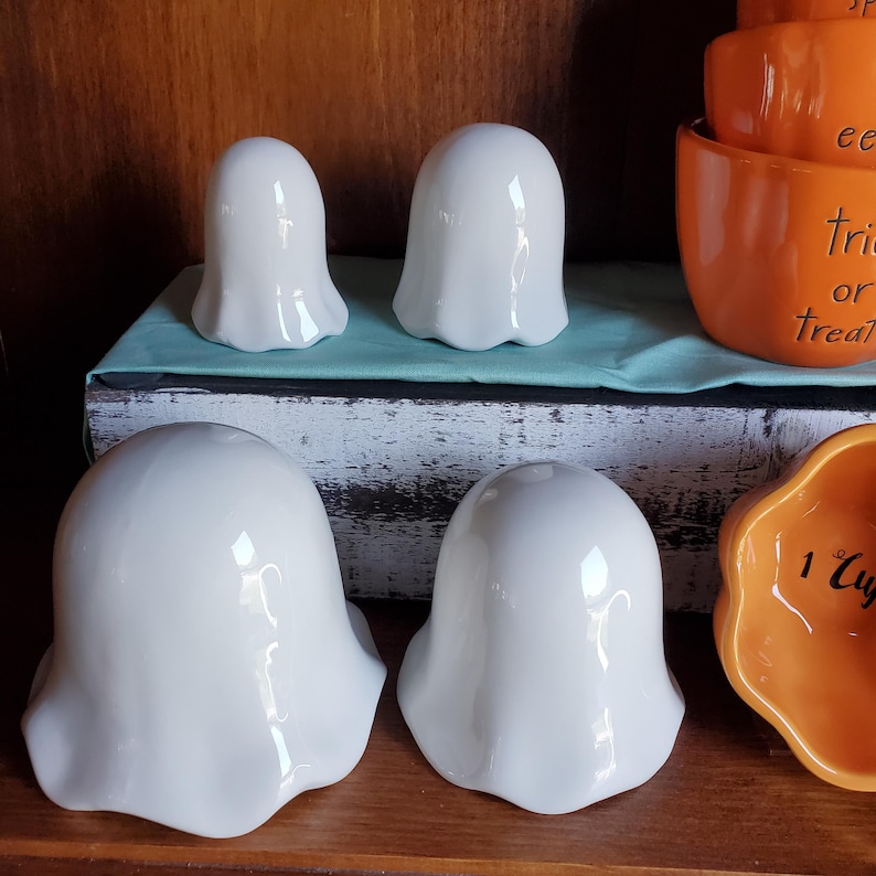 May include: Four white ceramic ghost-shaped measuring cups. The cups are different sizes and are arranged in a row on a wooden surface.