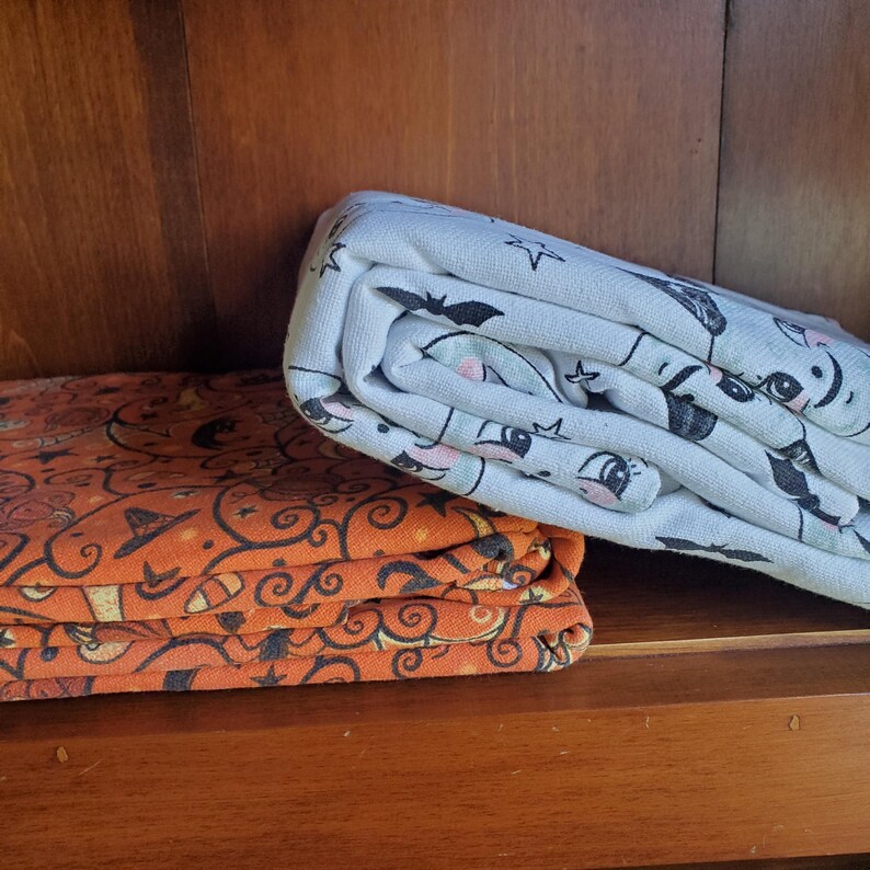 May include: Two folded fabric panels, one orange with a black and white Halloween-themed pattern and the other white with a black and white cartoon pattern.