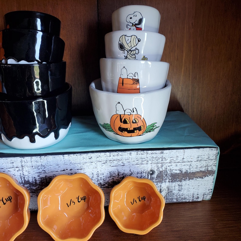 May include: Set of white ceramic measuring cups with Halloween-themed Peanuts characters. The largest cup features a pumpkin with a jack-o'-lantern face. The smaller cups feature Snoopy dressed as a mummy, a ghost, and a vampire. There are also three orange pumpkin-shaped measuring cups labeled 1/4 cup, 1/3 cup, and 1/2 cup.