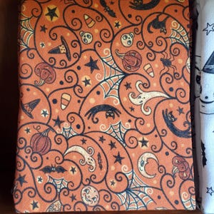 May include: Orange fabric with a black and white Halloween pattern. The pattern includes bats, spiders, pumpkins, ghosts, and stars.
