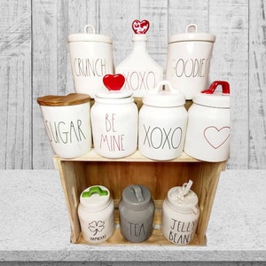 Puede incluir: Un conjunto de botes de cer&aacute;mica blanca con varias etiquetas, incluyendo "Sugar", "Crunch", "Goodies", "Be Mine", "XOXO" y "Jelly Beans". Los botes tienen acentos de coraz&oacute;n rojo y tr&eacute;bol y est&aacute;n dispuestos en un estante de madera.