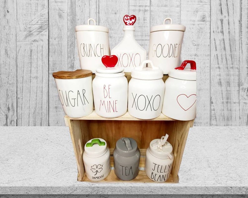 Puede incluir: Un conjunto de botes de cer&aacute;mica blanca con varias etiquetas, incluyendo "Sugar", "Crunch", "Goodies", "Be Mine", "XOXO" y "Jelly Beans". Los botes tienen acentos de coraz&oacute;n rojo y tr&eacute;bol y est&aacute;n dispuestos en un estante de madera.