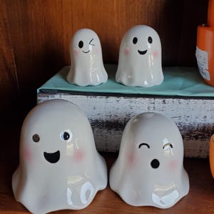 May include: Four white ceramic ghost figurines with different facial expressions. The ghosts have black eyes, mouths, and blush on their cheeks.