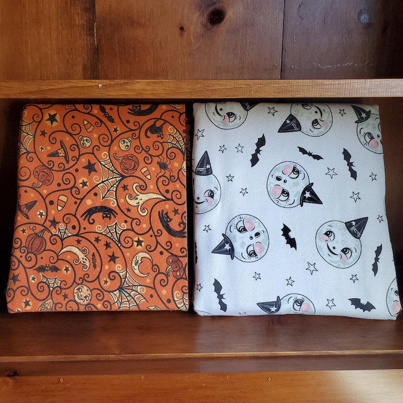 May include: Two fabric panels with Halloween-themed prints. One panel features a swirling orange and black pattern with ghosts, bats, and candy corn. The other panel is white with a repeating pattern of grey moons with faces and black bats.