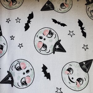 May include: White fabric with a repeating pattern of black bats, stars, and green witch faces with pink cheeks. The witch faces are wearing black witch hats.