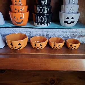May include: Set of four orange ceramic pumpkin-shaped bowls with black jack-o'-lantern faces. The bowls are arranged in a row from largest to smallest.