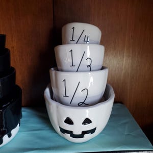 May include: A set of four white ceramic measuring cups with black markings. The cups are shaped like jack-o'-lanterns and are labeled 1/4, 1/3, 1/2, and 1 cup.