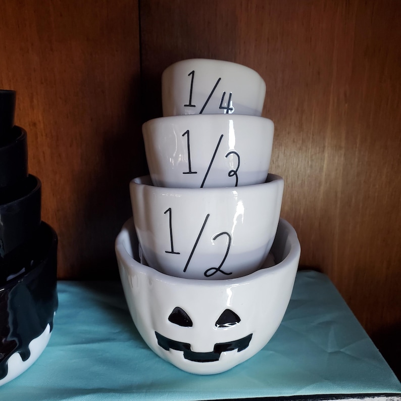 May include: A set of four white ceramic measuring cups with black markings. The cups are shaped like jack-o'-lanterns and are labeled 1/4, 1/3, 1/2, and 1 cup.