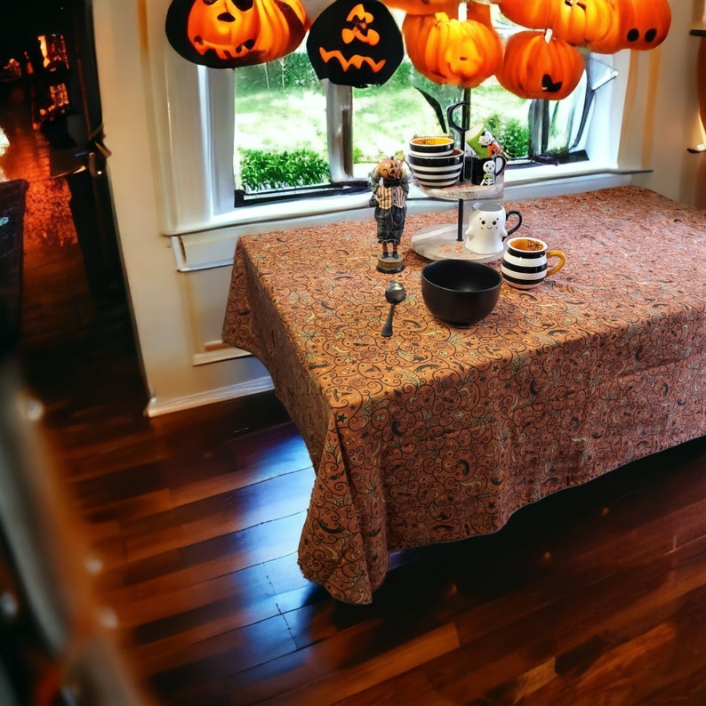 May include: A brown tablecloth with a black and white pattern of swirls and moons. A black bowl, a white mug with a black rim, and a white mug with a black ghost design are on the table. A small figurine of a ghost is on the table.