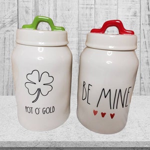 Puede incluir: Dos botes de cer&aacute;mica blanca con tapas verde y roja. El bote de la izquierda tiene un tr&eacute;bol de cuatro hojas y el texto "Pot O' Gold". El bote de la derecha tiene el texto "Be Mine" con tres corazones rojos.