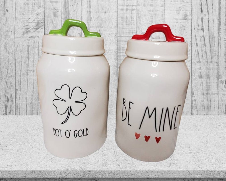 Puede incluir: Dos botes de cer&aacute;mica blanca con tapas verde y roja. El bote de la izquierda tiene un tr&eacute;bol de cuatro hojas y el texto "Pot O' Gold". El bote de la derecha tiene el texto "Be Mine" con tres corazones rojos.