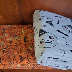 May include: Two Halloween-themed fabric panels. One panel features a white background with a repeating pattern of black bats, stars, and cartoon ghosts wearing witch hats. The other panel features an orange background with a repeating pattern of black bats, stars, and swirls with orange, black, and white accents.