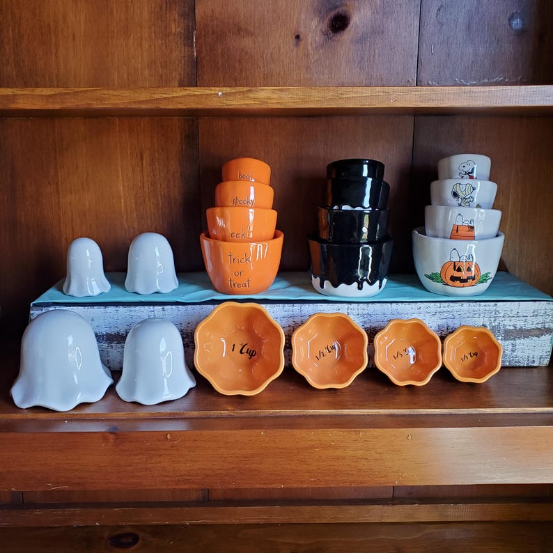May include: A set of orange and black Halloween themed measuring cups and bowls. The cups are labeled with measurements: 1 cup, 1/2 cup, 1/3 cup, and 1/4 cup. The bowls are labeled with the text "Trick or Treat". There are also two white ghost-shaped bowls.