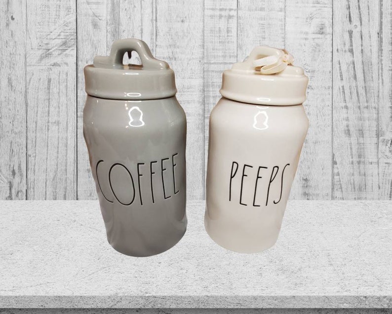 Puede incluir: Dos botes de cer&aacute;mica con tapas, uno gris con la palabra "COFFEE" y otro blanco con la palabra "PEEPS".