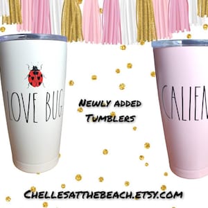 Puede incluir: Dos vasos térmicos de acero inoxidable con tapas. El vaso blanco tiene una mariquita roja y el texto "LOVE BUG". El vaso rosa tiene el texto "CALLIENTE".