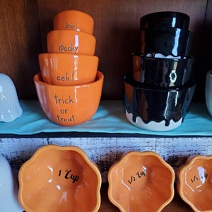 May include: Set of orange and black Halloween themed measuring cups. The orange cups are shaped like pumpkins and have the words "boo!", "spooky", "eek!", "trick or treat", "1 cup", "1/2 cup", and "1/3 cup" written on them. The black cups are stacked and have a dripping black glaze.