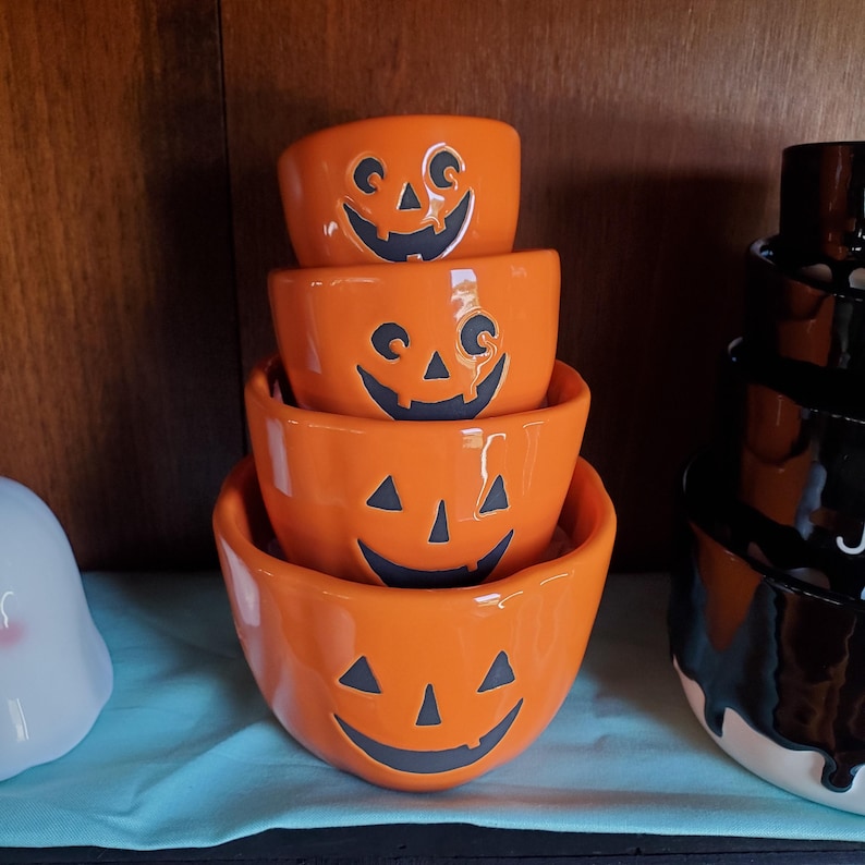 May include: Set of four orange ceramic pumpkin-shaped bowls with black jack-o'-lantern faces. The bowls are stacked in order of size, with the largest bowl at the bottom and the smallest bowl at the top.