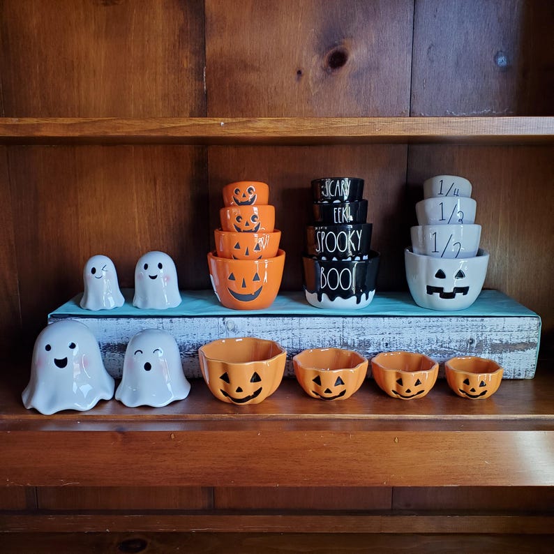 May include: A set of Halloween-themed ceramic measuring cups and bowls. The cups are white with black lettering and feature the phrases "1/4", "1/3", and "1/2". The bowls are orange and black with a pumpkin face design. There are also two white ghost-shaped bowls and a set of four smaller orange pumpkin-shaped bowls.