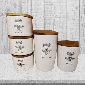 Puede incluir: Conjunto de cuatro botes de cer&aacute;mica blanca con tapas de madera. Cada bote tiene una ilustraci&oacute;n de abeja en blanco y negro y una frase: "queen", "bee happy", "bee kind", y "queen".