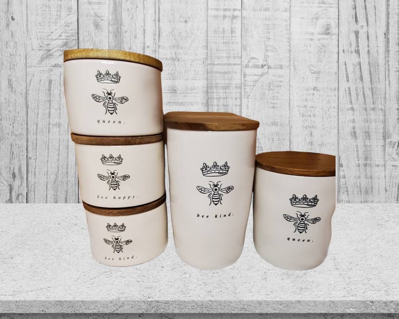 Puede incluir: Conjunto de cuatro botes de cer&aacute;mica blanca con tapas de madera. Cada bote tiene una ilustraci&oacute;n de abeja en blanco y negro y una frase: "queen", "bee happy", "bee kind", y "queen".